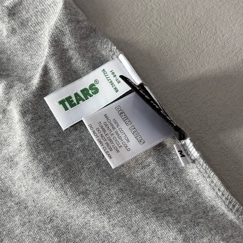 Close-up of tags on a Denim Tears Grey Cotton Wreath Allover Print T-Shirt - 2 in heather grey. Tags feature TEARS® in green and DENIM TEARS, along with sizing, material, and care info printed in black.