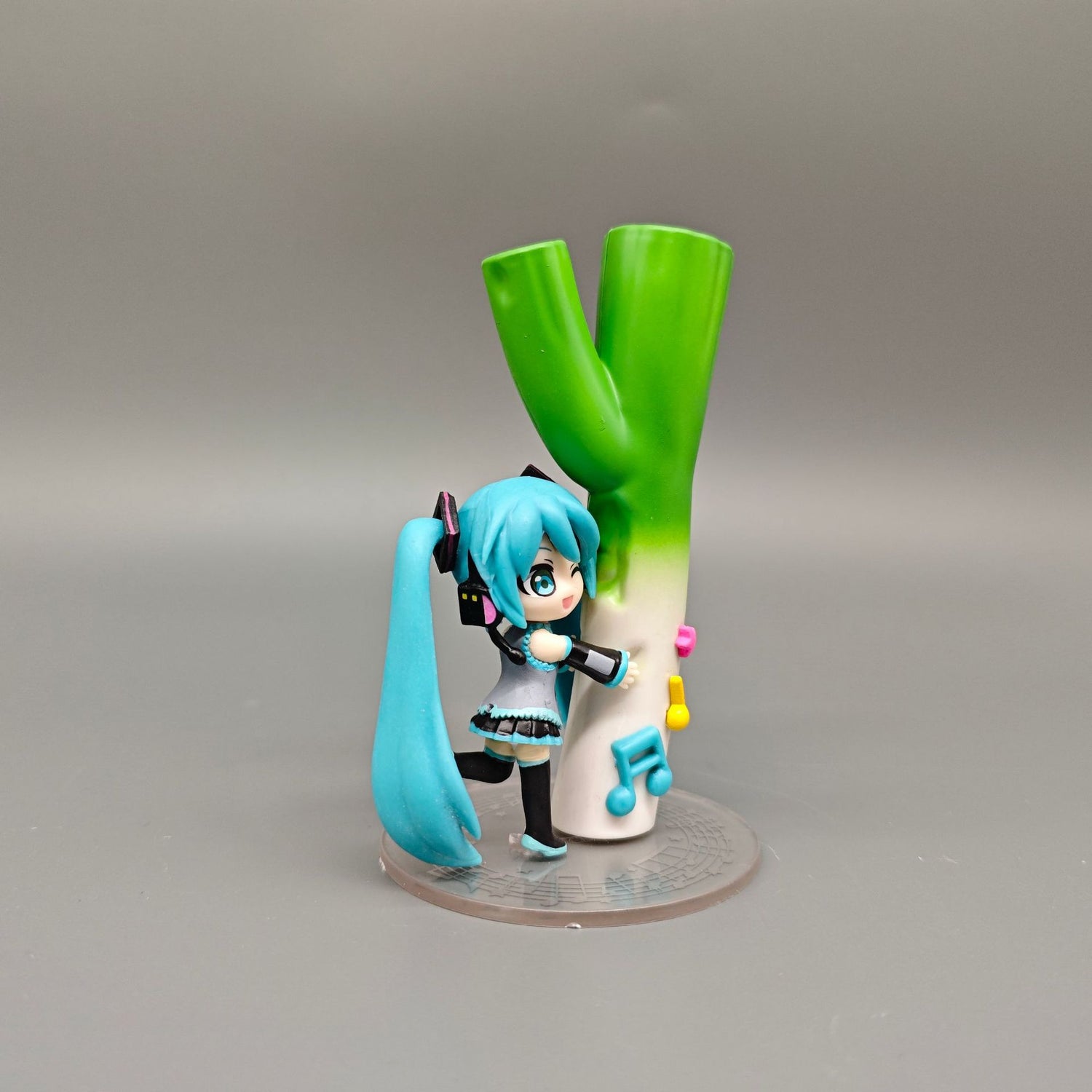 The Chibi Hatsune Miku 12cm PVC Figure by Hatsune Miku features a playful figure with long blue hair holding a green onion, adorned with musical notes and bells, set against a plain gray background.