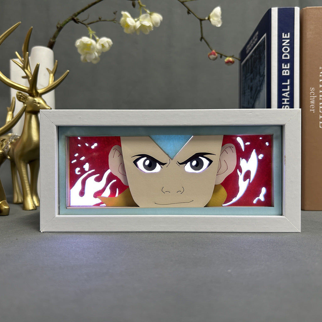 A framed image of an animated character in the Avatar State, with a glowing arrow and red flames, sits beside an Aang Avatar State LED Light Box – Avatar: The Last Airbender 3D Lamp by Avatar the Last Airbender, against a backdrop of books, white flowers, and a golden deer figurine.