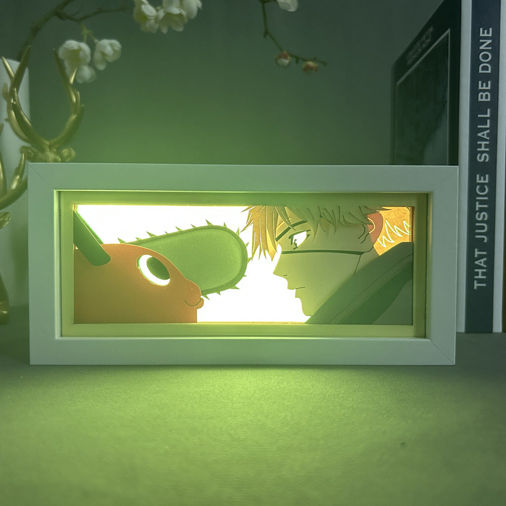 The Chainsaw Man Anime Light Box – Denji &amp; Pochita 3D LED Lamp by Chainsaw Man displays a dynamic illustration of Denji with spiky hair and Pochita, featuring color-changing effects against a greenish glow, subtly revealing a book and vase of flowers in the background.