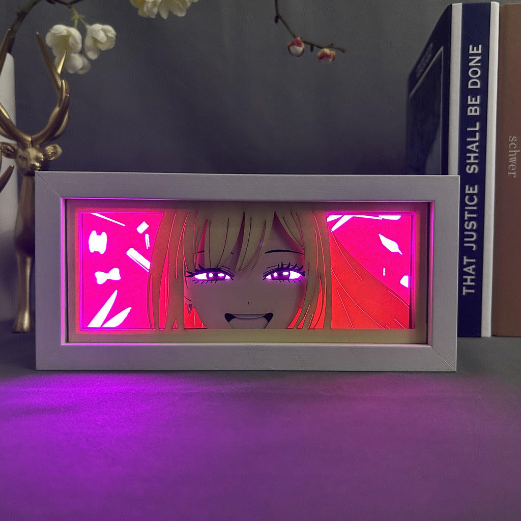 A framed artwork depicting an anime-style illustration of a character with long hair and bright eyes on a vibrant pink background resembles the "Chainsaw Man Anime Light Box – Power Smiling 3D LED Lamp with Dynamic Color-Changing Effects," displayed on a table with decorative objects and books.