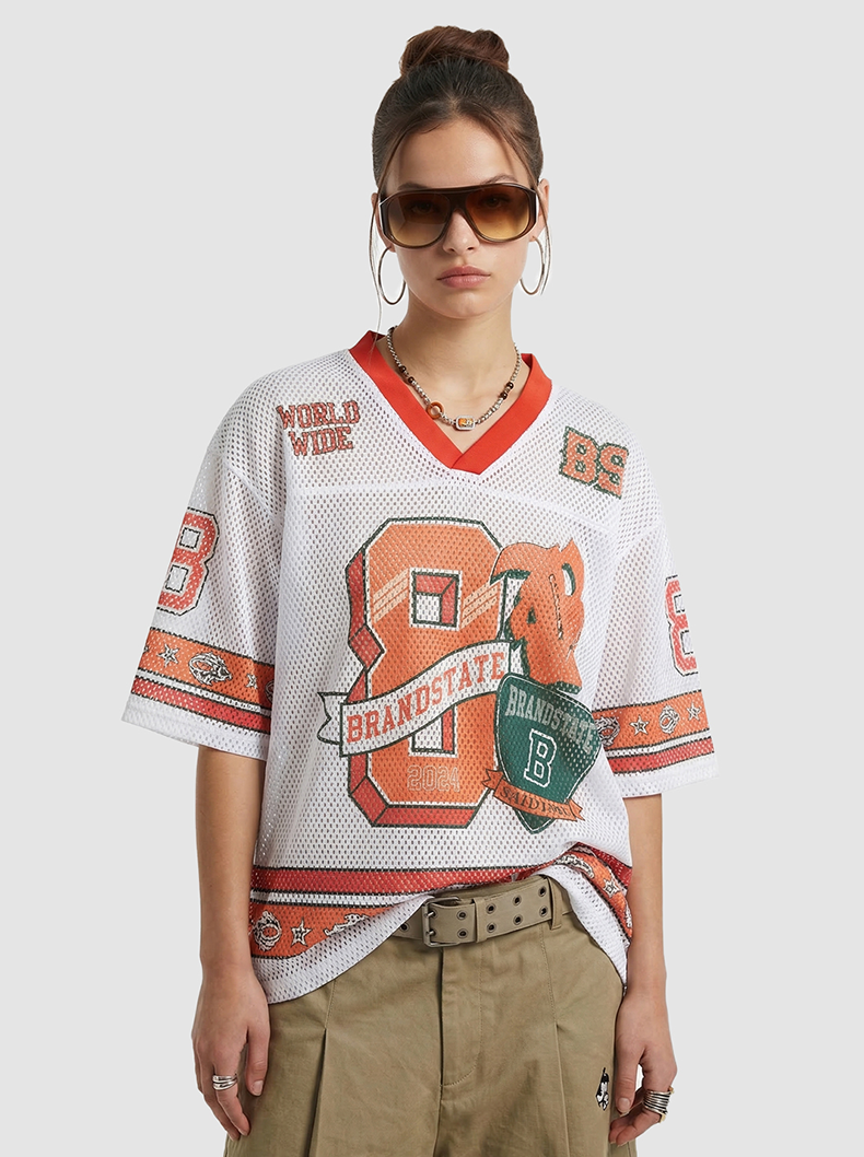 Model wearing oversized white mesh varsity jersey T-shirt with orange and green graphic print and red V-neck trim