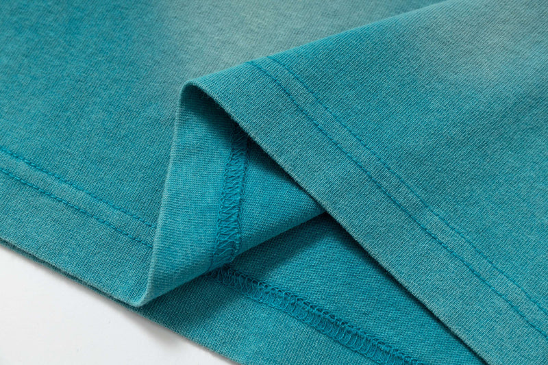 Close-up of a teal washed cotton tee hem with double stitching and raw edges, showing vintage streetwear texture