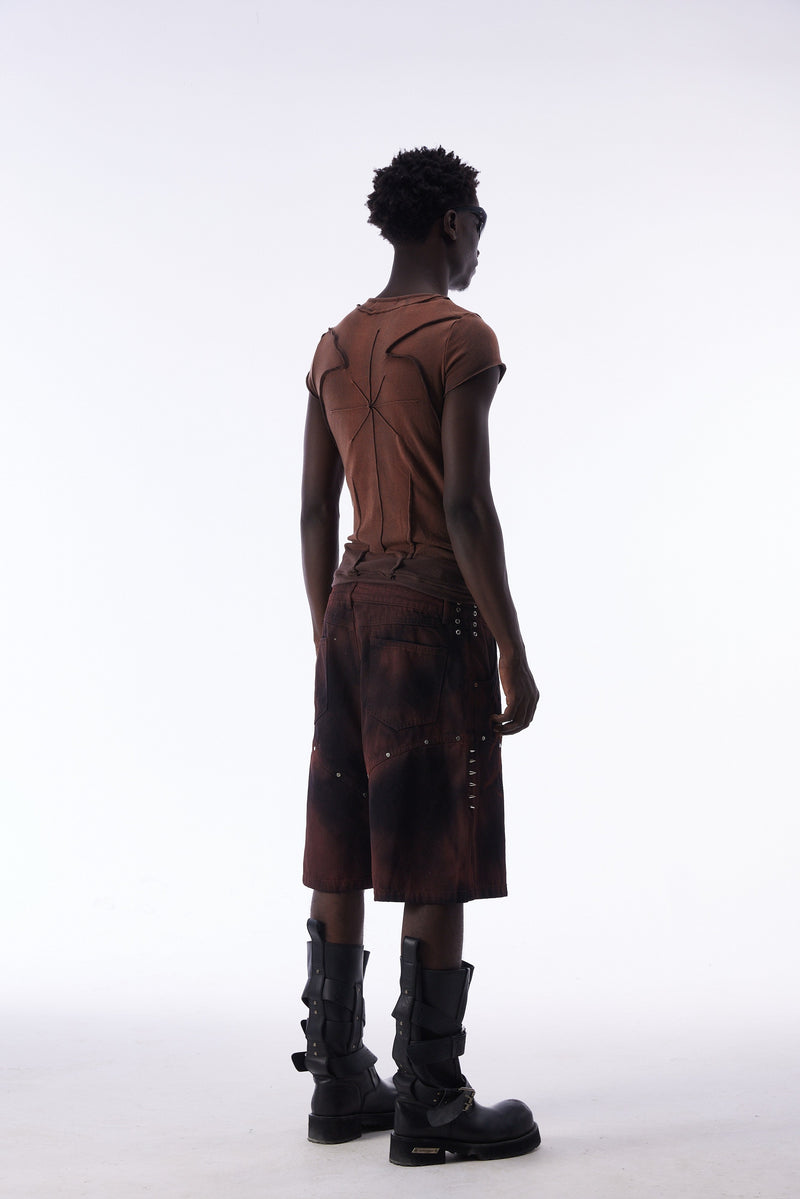 Model wearing a sculpted brown raw-seam cotton tee with dark panel shorts and black buckled boots on a white background