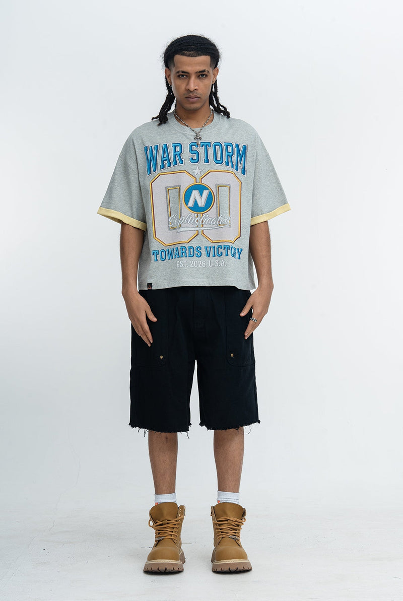 Model wearing gray Seakoff War Storm varsity graphic tee with blue lettering, black shorts, and tan boots