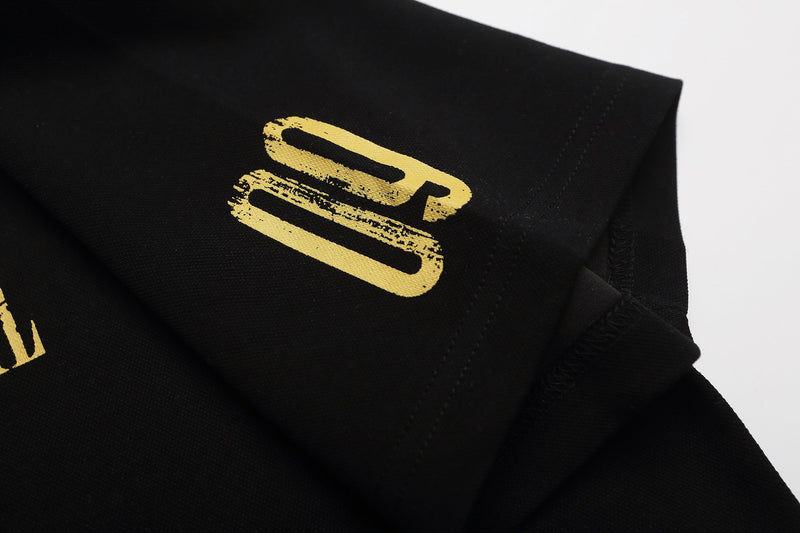 Black graphic polo shirt with gold distressed print and folded collar detail on a clean white background