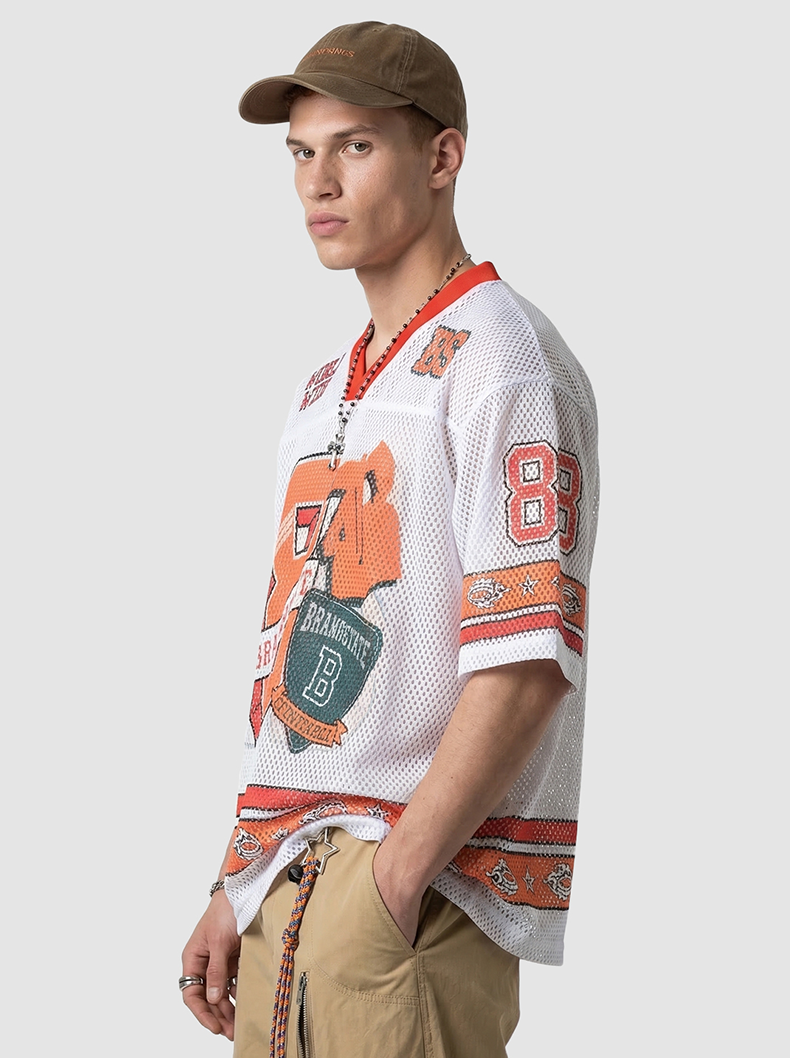 Model wearing a white mesh varsity jersey with orange graphics, red trim, khaki pants, and a brown cap on a studio background