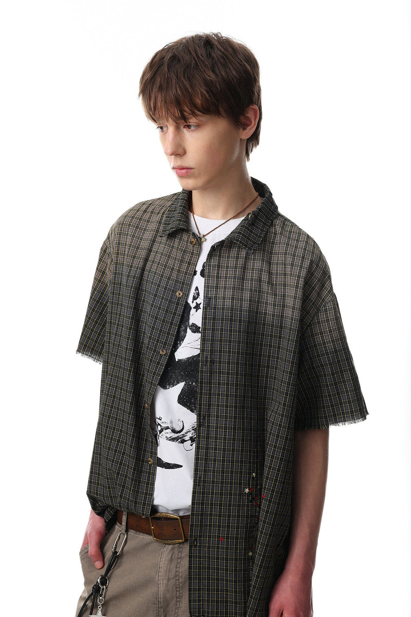 Model wearing a dark gradient plaid short-sleeve shirt with raw edges over a graphic tee, styled with khaki pants