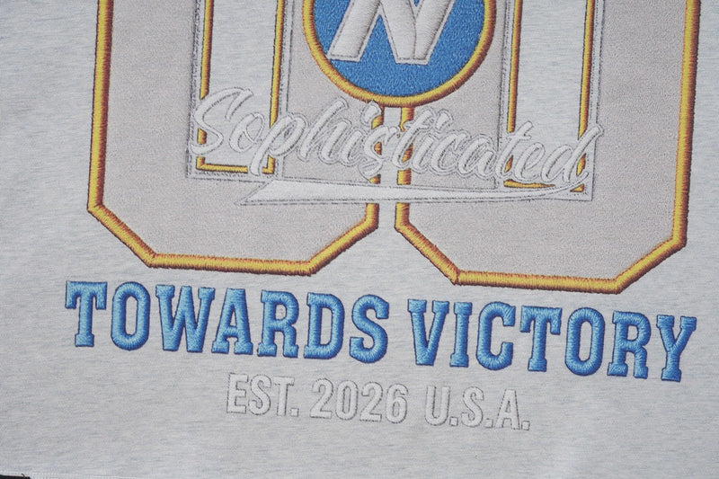 Close-up of a white varsity graphic tee with blue Towards Victory lettering, gold outlined emblem, and Est. 2026 U.S.A. print