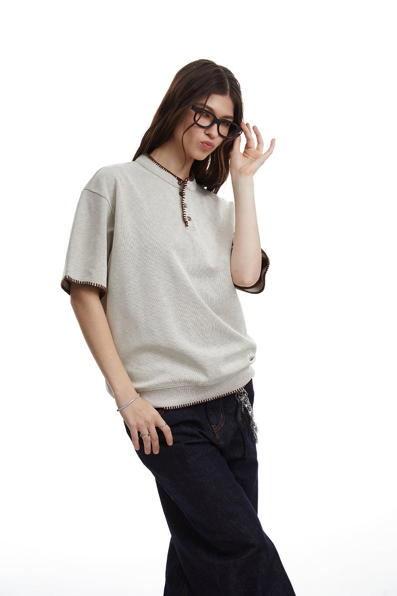 Relaxed-fit beige Henley tee with contrast stitching, worn with dark jeans and sunglasses on a white background