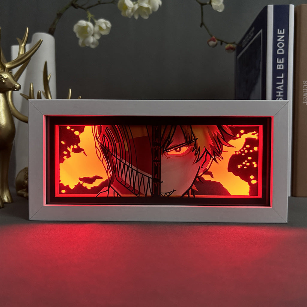 A rectangular frame showcases a Chainsaw Man Anime Light Box, featuring Denji in his chainsaw form. Its split-face design contrasts human and robotic elements with dynamic color-changing effects, enhanced by a fiery red background. Nearby are books, a white vase, and a golden deer figurine.