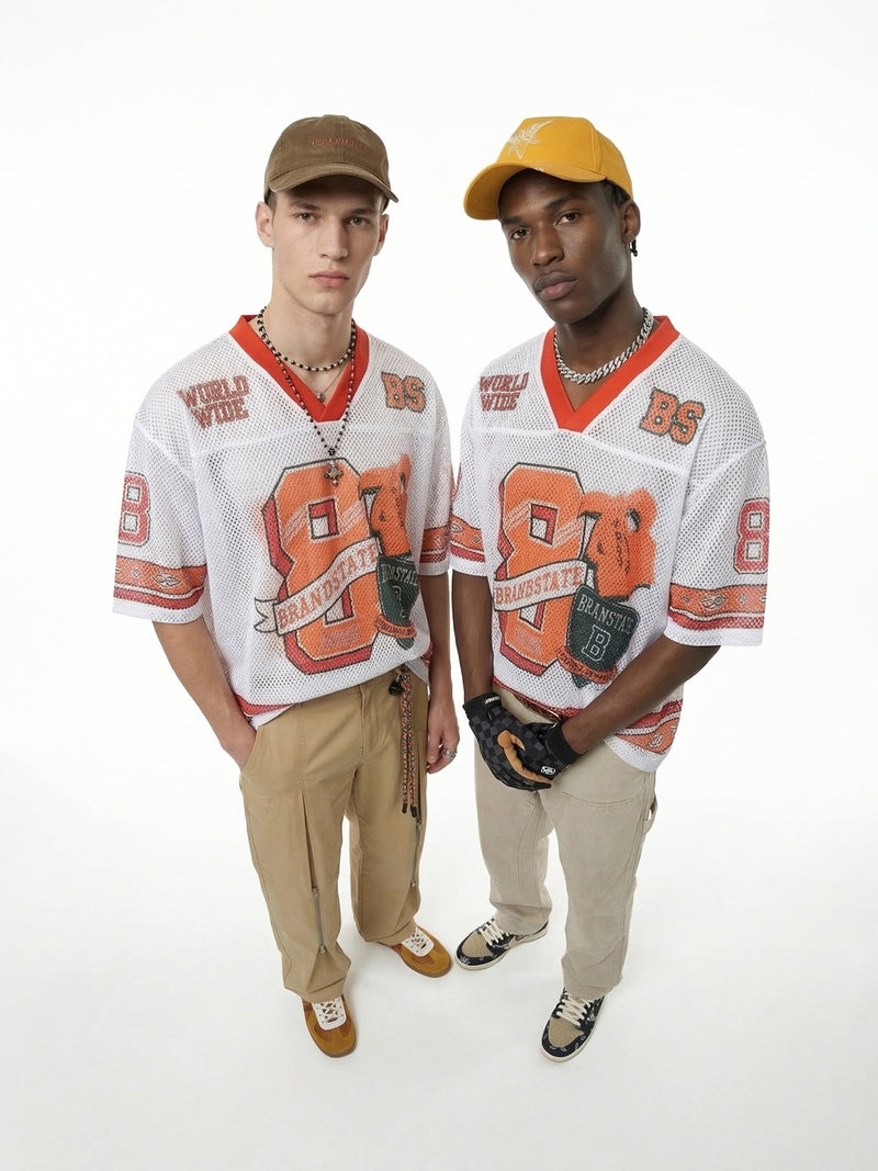 Two models wearing oversized white mesh varsity jerseys with orange graphics, caps, and beige pants on a white background
