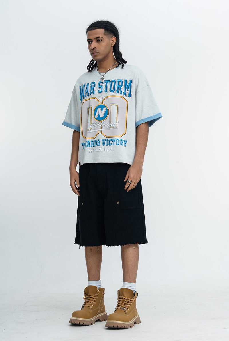 Model wearing an oversized light gray War Storm varsity graphic tee with blue trim, styled with black shorts and tan boots