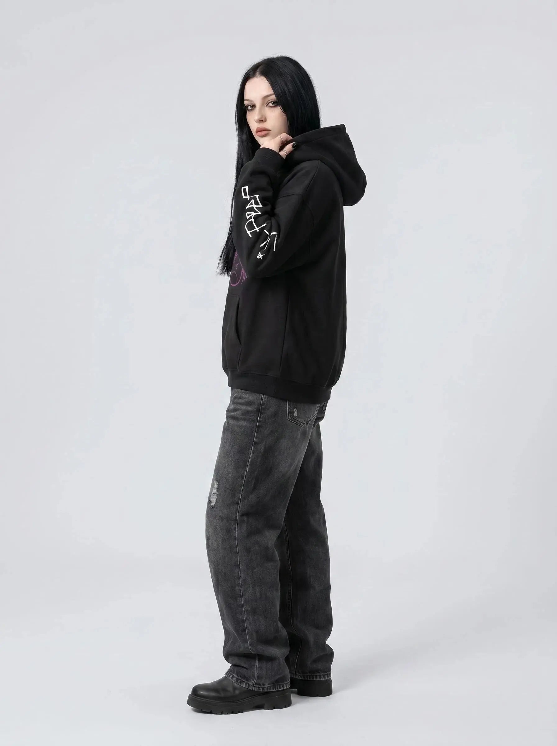 A person with long black hair wears the Lil Peep Hoodie — Sad Face Graffiti Graphic Sweatshirt, paired with loose dark jeans and black shoes, standing against a plain light grey background.