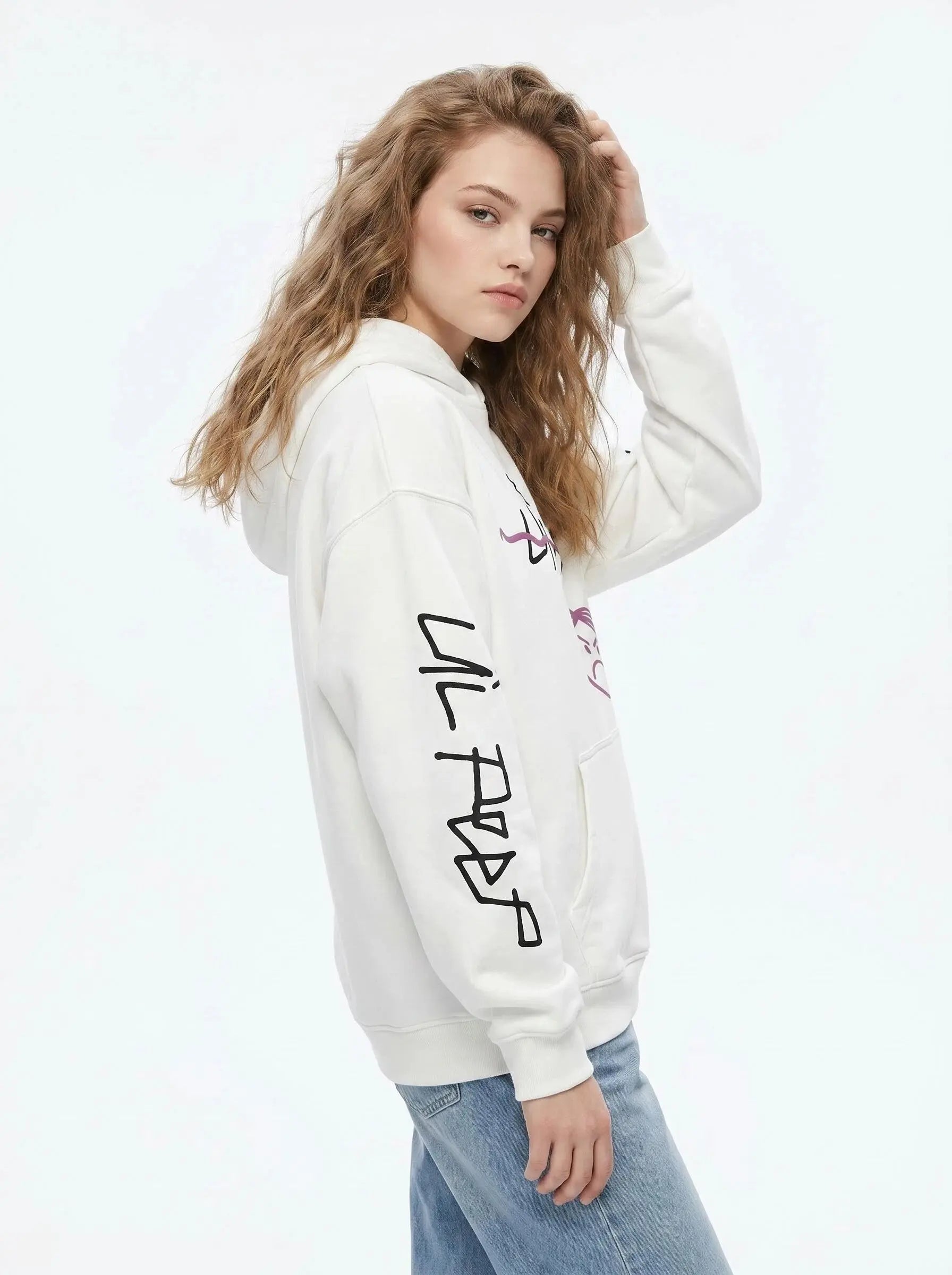 A person with wavy light brown hair wears the Lil Peep Hoodie — Sad Face Graffiti Graphic Sweatshirt, featuring black graphics on the sleeve, along with blue jeans, standing against a plain white background and gazing at the camera.