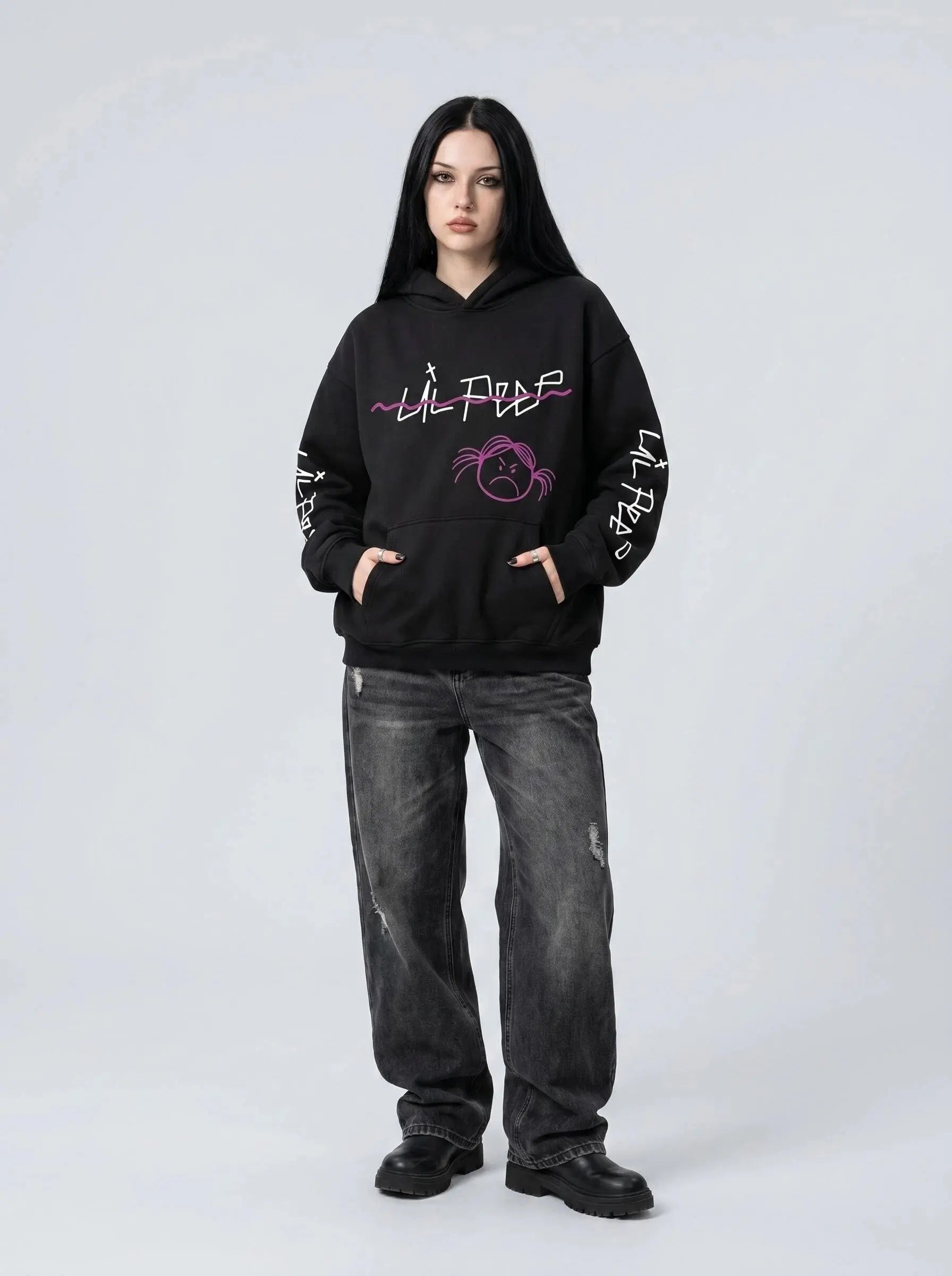 A person with long dark hair in a black Lil Peep Hoodie — Sad Face Graffiti Graphic Sweatshirt, loose dark jeans, and black boots stands against a plain light gray background.