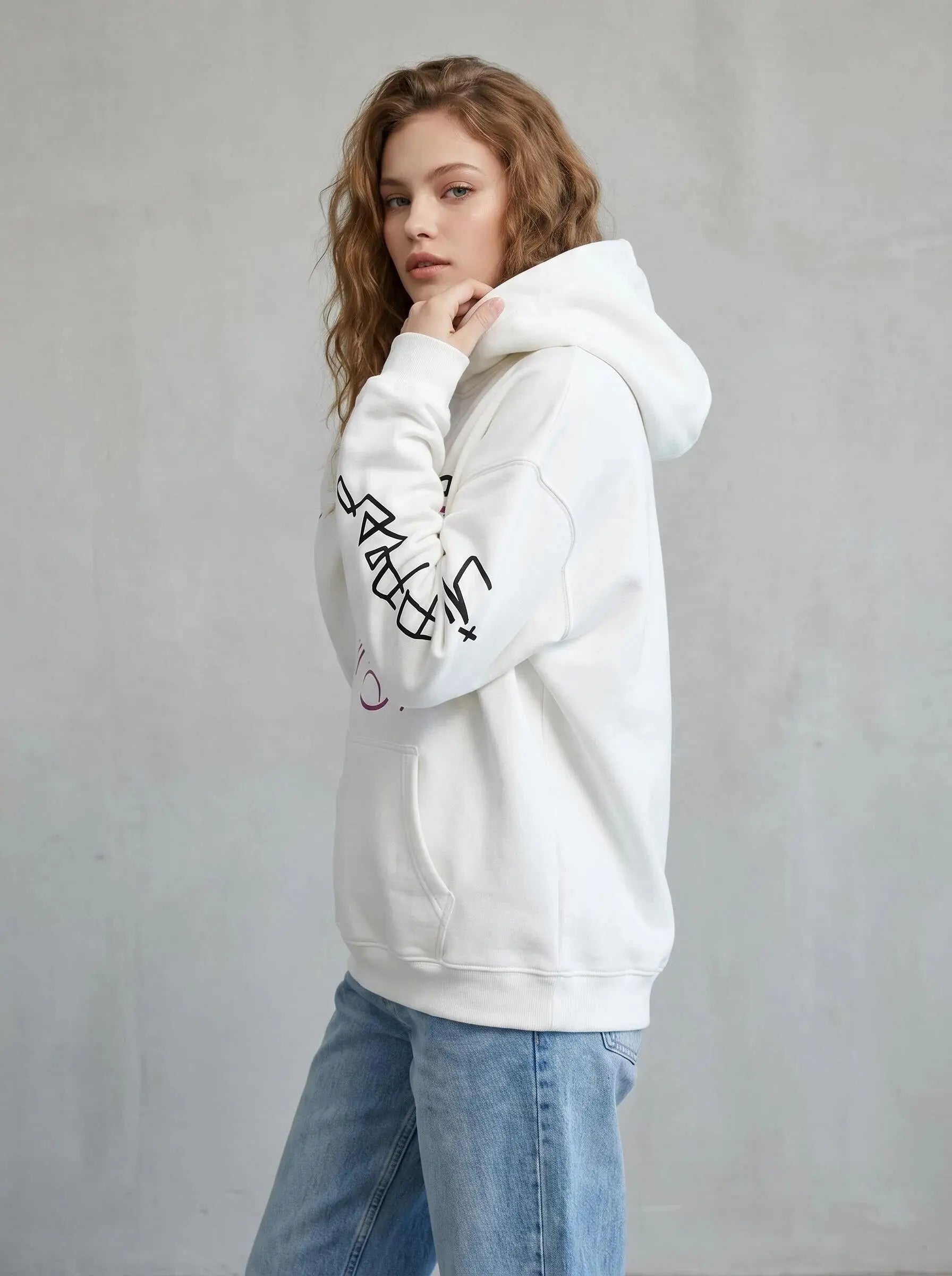 A young woman with wavy, light brown hair models the white Lil Peep Hoodie — Sad Face Graffiti Graphic Sweatshirt with black sleeve graphics and light blue jeans, posing sideways against a plain gray background.