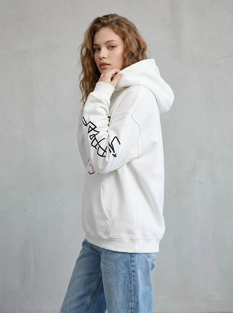 A young woman with wavy, light brown hair models the white Lil Peep Hoodie — Sad Face Graffiti Graphic Sweatshirt with black sleeve graphics and light blue jeans, posing sideways against a plain gray background.