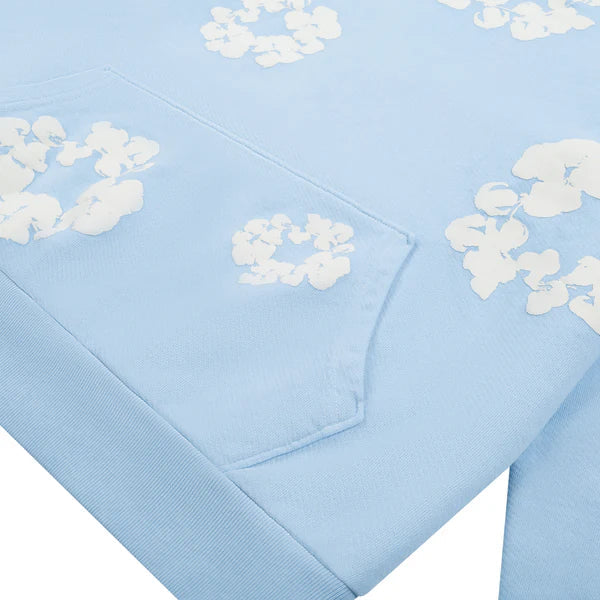 The Light Blue Denim Tears Hoodie – White Cotton features white floral prints and a visible pocket on its light blue fabric, displayed flat to showcase its stylish, comfortable design.