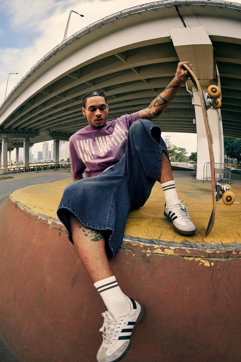 Model in dark wide-leg denim shorts, side view, seated with skateboard beside him