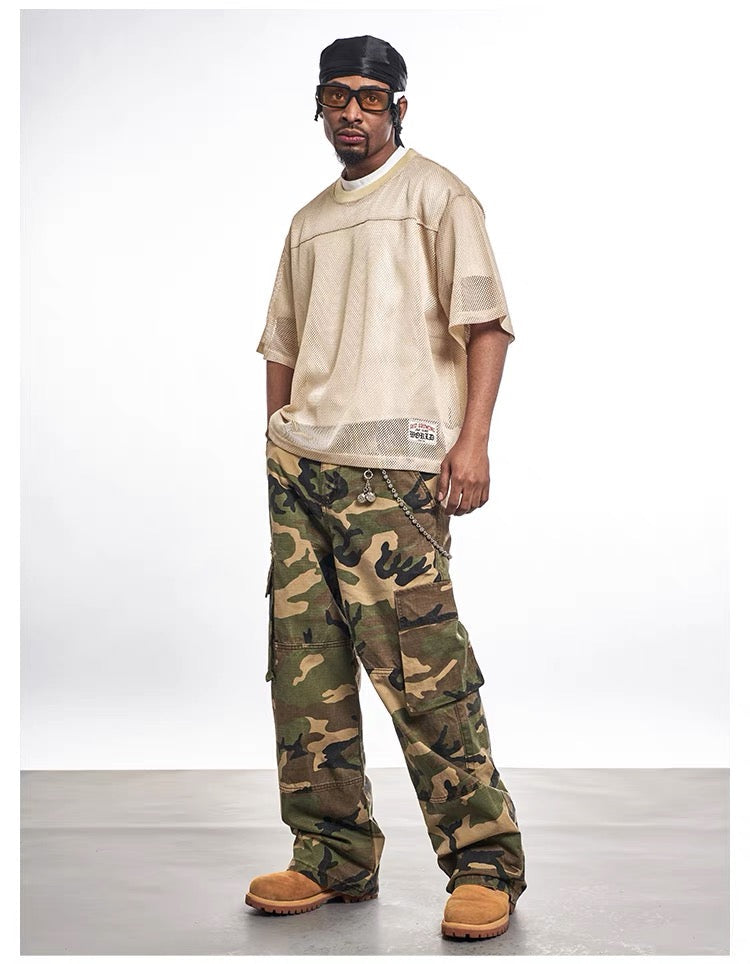 Model wearing a beige mesh jersey tee with camo cargo pants and tan boots in a streetwear studio shot