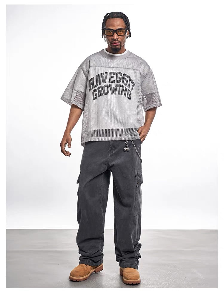 Model wearing a gray mesh varsity graphic tee with loose black cargo pants and tan boots on a white studio background