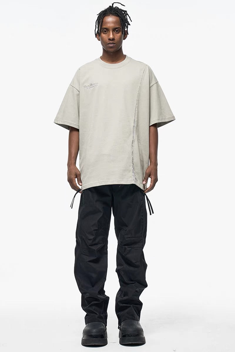 Model wearing an oversized beige asymmetric stitch tee with raw seams, paired with black cargo pants and boots