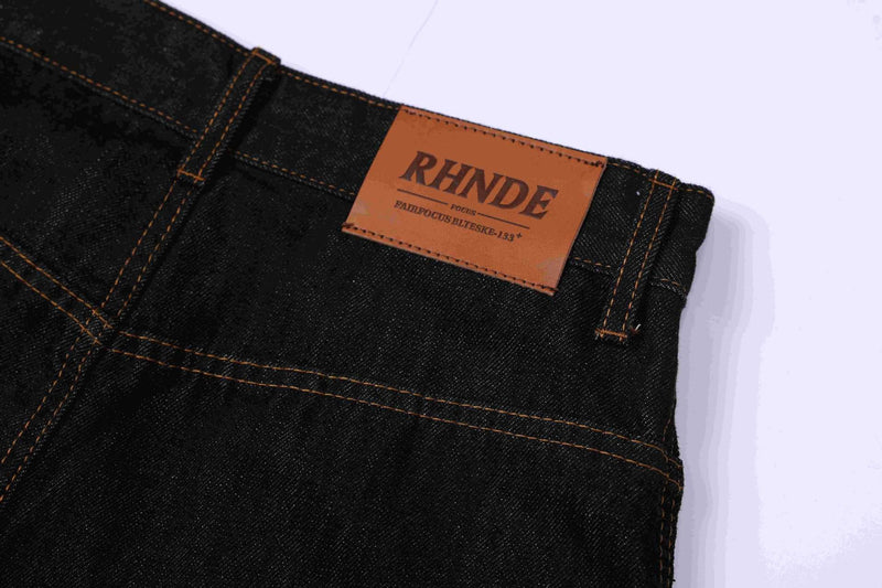 Close-up of dark indigo denim jorts waistband with leather brand patch and orange stitching