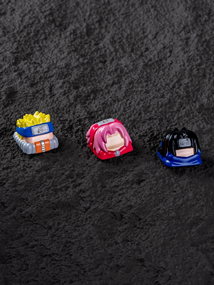 Three anime-esque ninja figurines resembling the Naruto Keycap Set are set on a dark surface. Each features a unique hairstyle and headband: one yellow, one pink, and one black—ideal companions for mechanical keyboard enthusiasts. Product: Team 7 Naruto Keycap Set by Naruto.