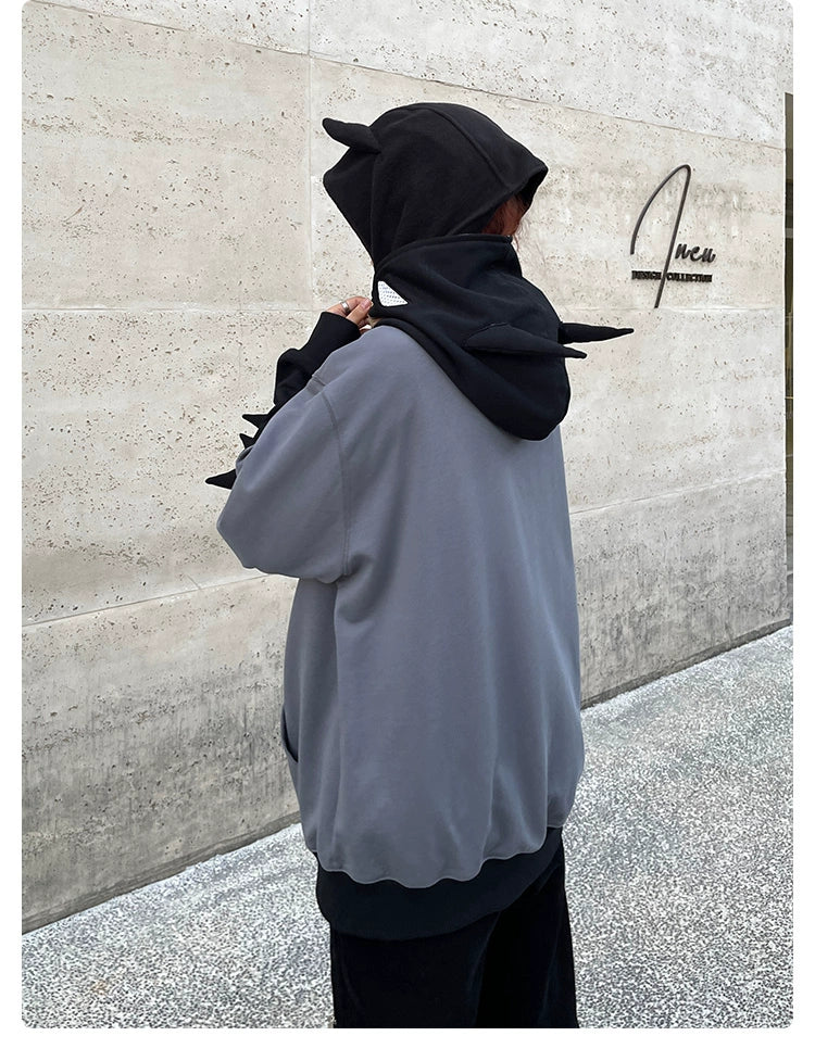 A person wearing a Seakoff Batman-Inspired Hoodie—a grey and black 400GSM oversized zip-up with cat ears and tail—stands before a textured beige wall with a sign. With their back to the camera, this superhero streetwear piece adds intrigue to the scene.