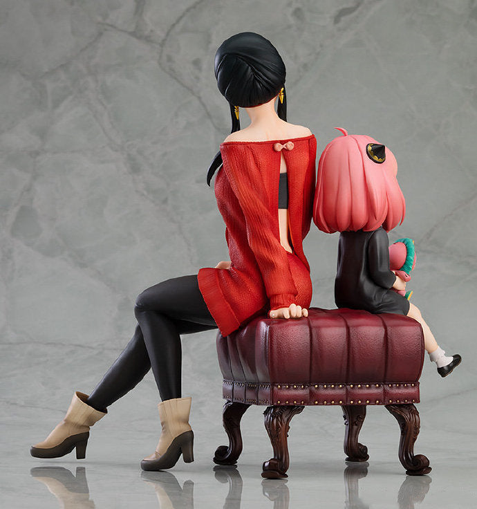 The Spy × Family Anya and Yor PVC Figure set captures an adorable mother-daughter moment with Yor, in a red outfit with beige boots sitting on a tufted brown bench, and Anya, in a black dress holding a green object.