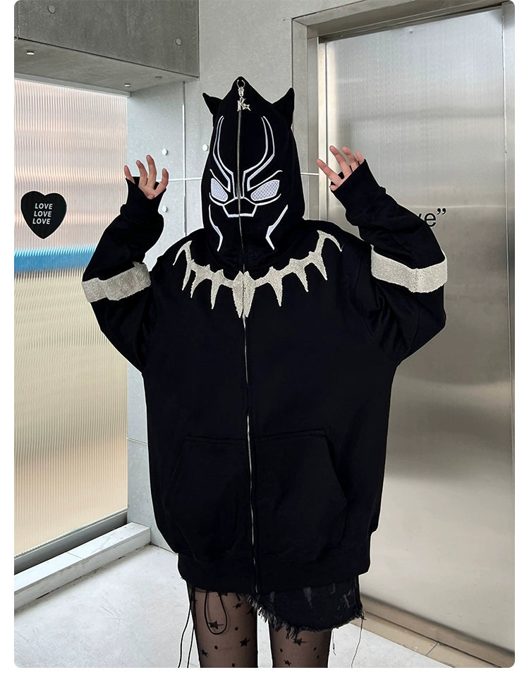 Indoors, a person wears a Seakoff Black Panther-Inspired Hoodie—a 400GSM oversized superhero zip-up sweatshirt with an ear design. Silver details highlight their black gloves. The hoodie covers their face, and paired with patterned tights and a black skirt, it appeals to streetwear fans.