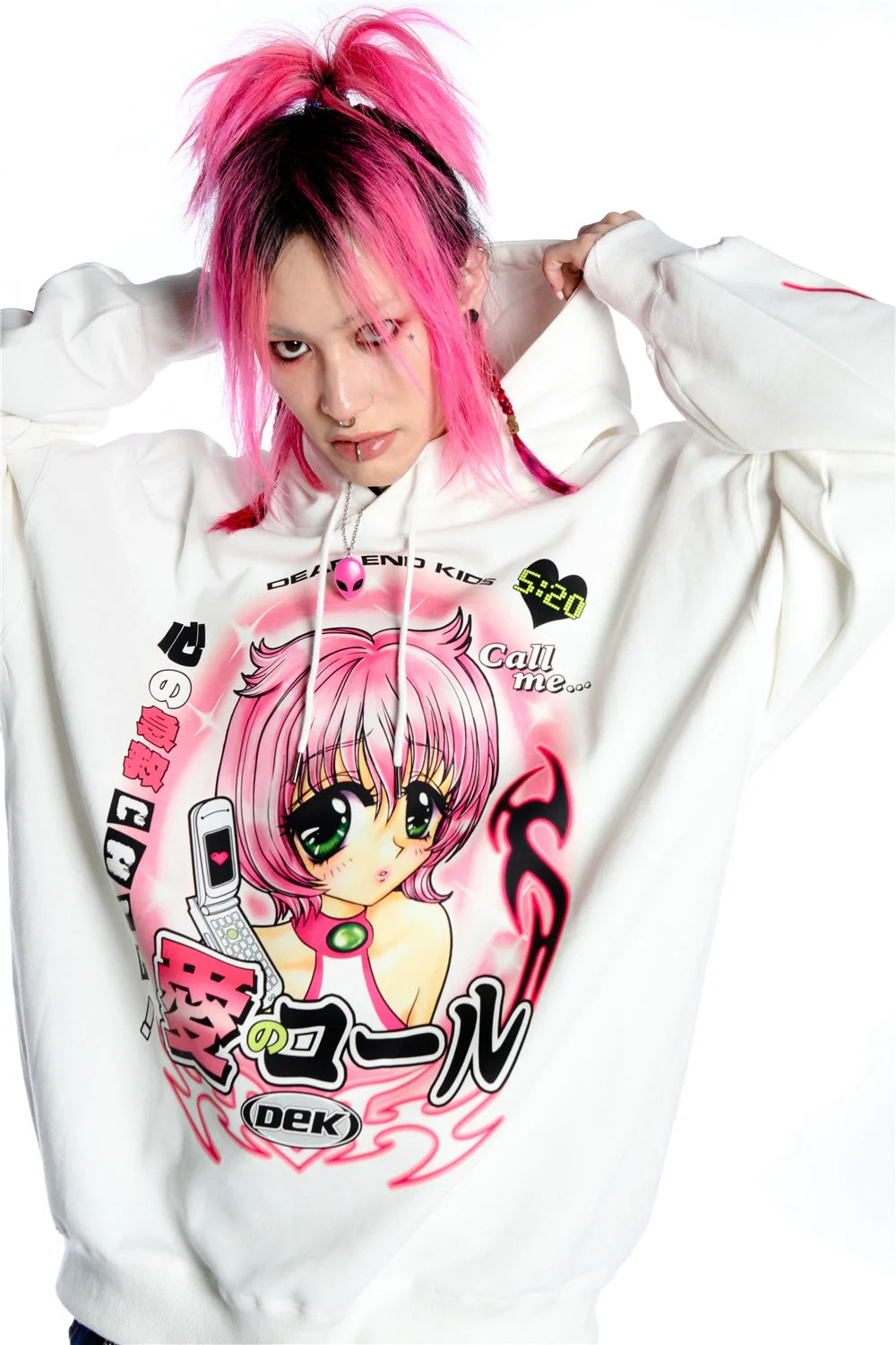 With pink hair in a high ponytail, they epitomize the kawaii aesthetic wearing Seakoff's Y2K Aesthetic Anime Hoodie, adorned with a retro manga girl graphic holding a phone. Posing hands behind their head against a plain background, they capture the playful Y2K vibe.