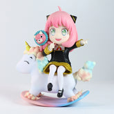 The Spy x Family Anya Forger PVC Figure (15 cm) portrays Anya with pink hair and cat-like ears, dressed in a black dress with gold accents, sitting on a pastel unicorn rocking horse. Featuring an interchangeable face expression, it embodies charm alongside her tiny doll.