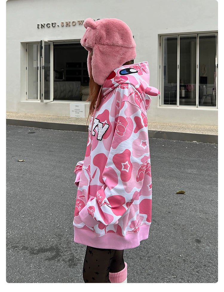 Dressed in a Seakoff Pink Kirby-Inspired Hoodie, featuring 400GSM cotton, camo design, and 3D ears, the person pairs it with a pink bear hat while standing on gray pavement. A white building with &quot;INCU. SHOW&quot; above a window contrasts with their playful outfit.