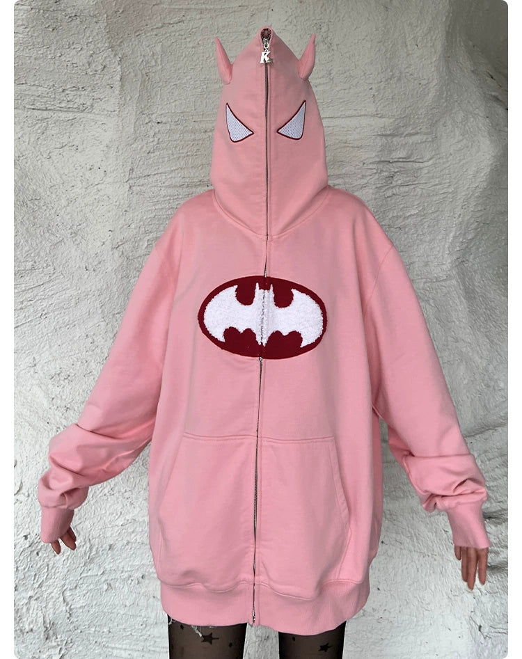 A person wears the oversized Batman-Inspired Hoodie from Seakoff, a pink zip-up sweatshirt with pointed ears on the hood and a white and maroon bat-like symbol on the front. It covers their face as they stand by a light wall, creating an ideal look for superhero fans.