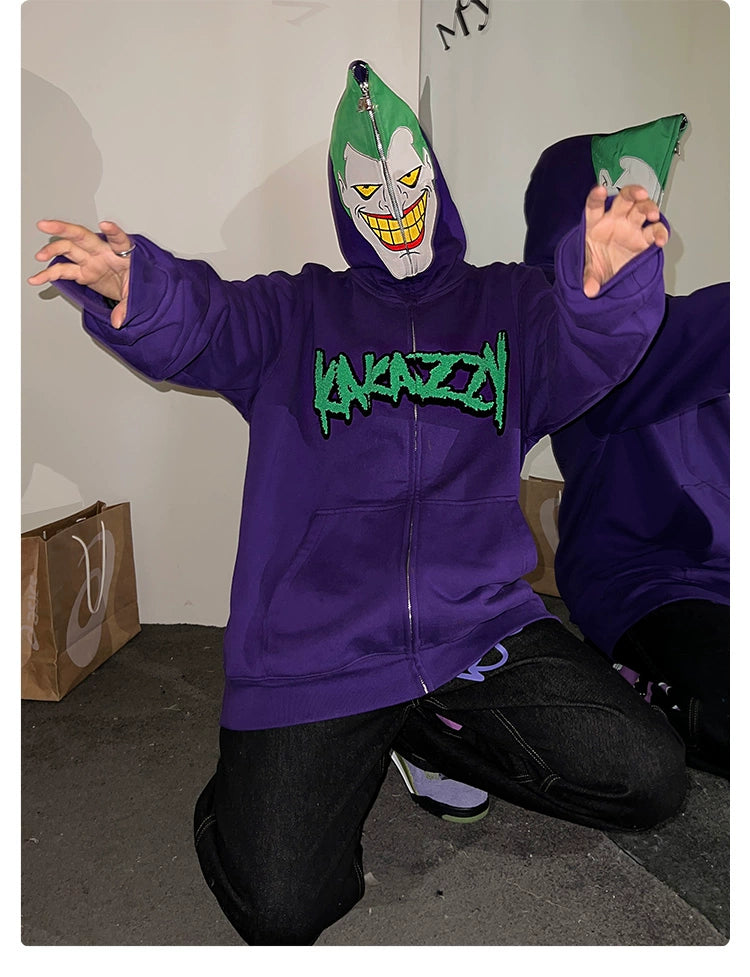 Kneeling with outstretched hands, a person wears the Joker-Inspired Hoodie by Seakoff—a 400GSM oversized purple zip-up sweatshirt. Its green lettering pairs with a grin-adorned green-and-white mask, while behind them sits a shopping bag ideal for supervillain fans.