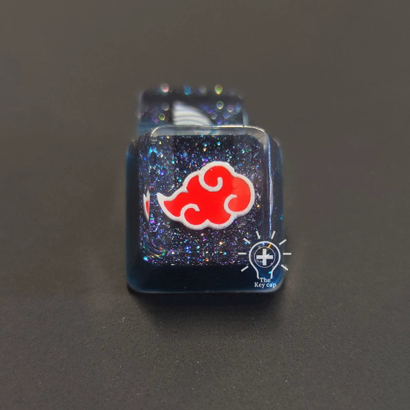 A premium Naruto Symbols Keycap Set offers a resin black mechanical keyboard keycap with shimmering blue and purple hues, featuring a red cloud design outlined in white. It creates an illuminated effect against its dark backdrop on a black surface.