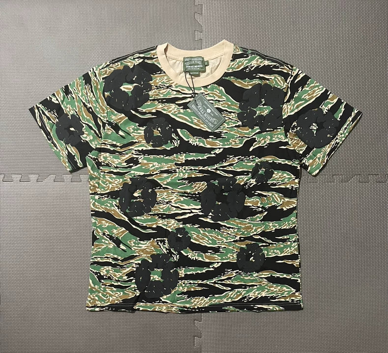 The Denim Tears Green Camo All-Over Print T-Shirt features short sleeves, a green, brown, and black camouflage pattern with bold black flower prints, cotton fabric, attached tags, and is shown on a gray textured surface.