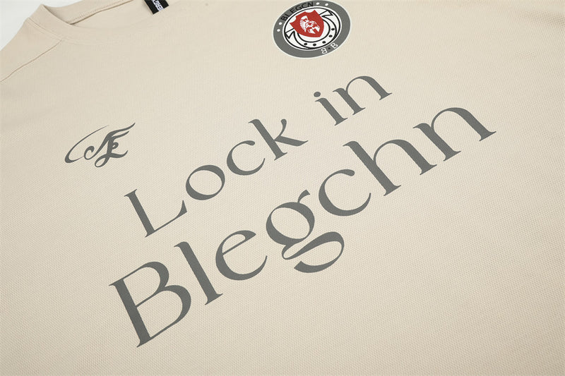 Beige tee front print detail with chest logo, crest patch, and large Lock in Blegchn text