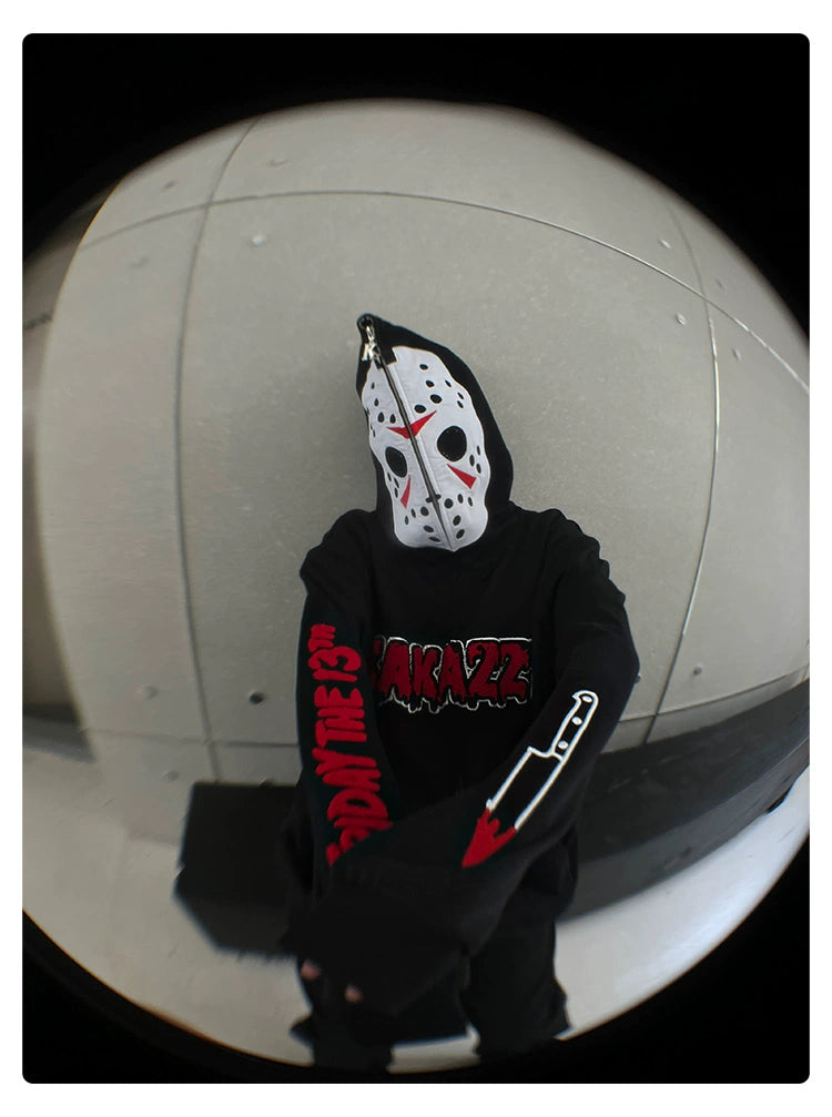 In a fisheye lens photo, a person wearing a Seakoff Friday the 13th Hoodie and a Jason mask poses against a concrete wall. This oversized zip-up sweatshirt features red and white designs, text, and a knife graphic, making it ideal for horror movie fans.