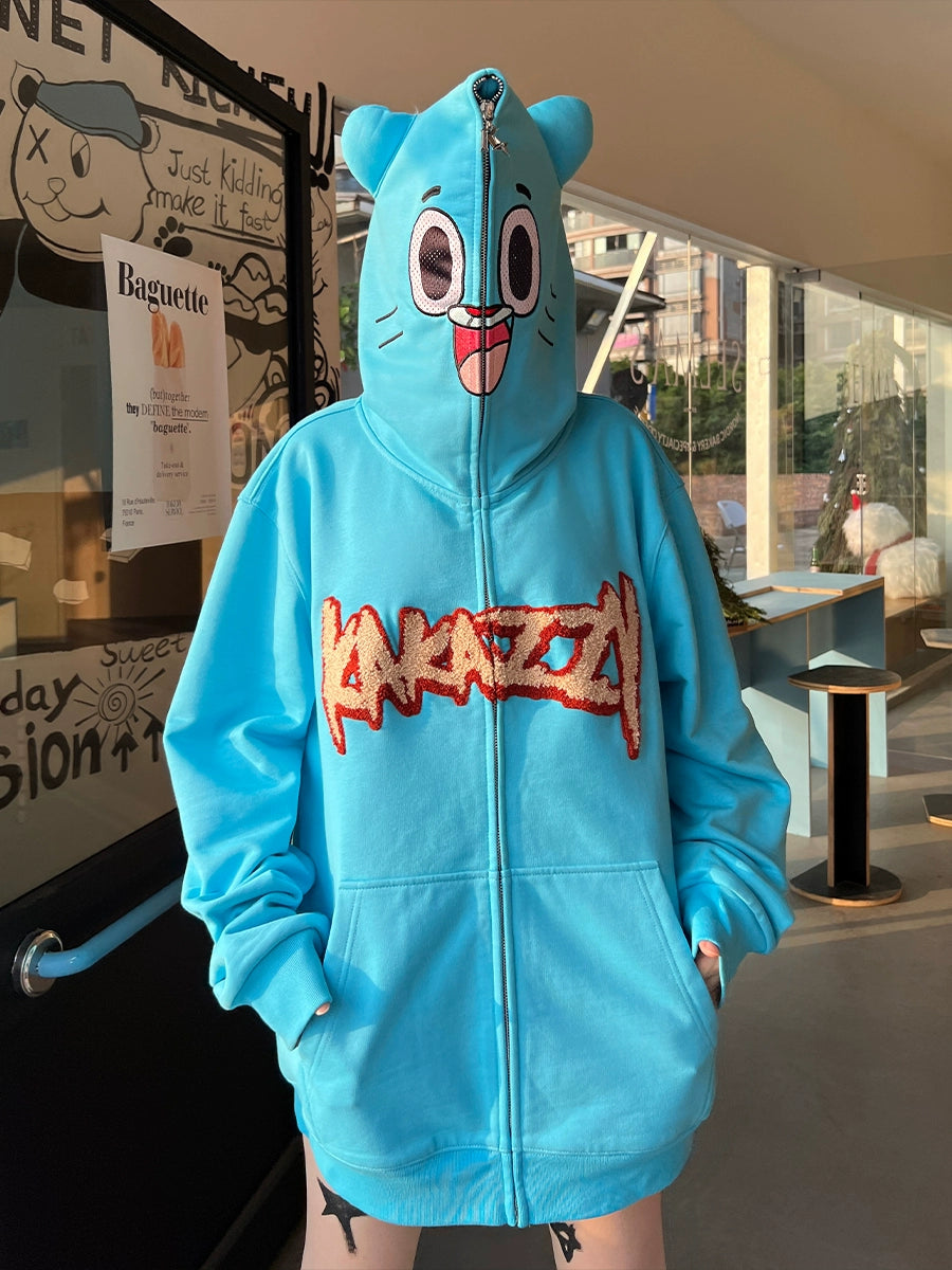 A person wears a Seakoff Adorable Blue Cartoon Hoodie made of 400GSM cotton, featuring playful eyes and ears on the hood. The front is stylized with &quot;ANAZIZM&quot; in red. Through a glass door, a wall poster complements the cartoonish vibe.