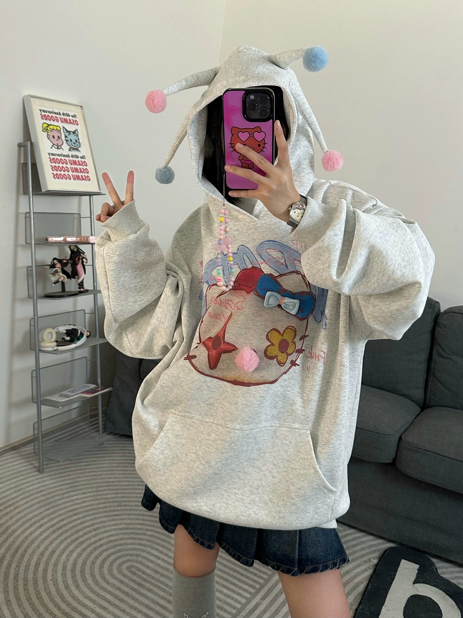 Wearing a Seakoff Kawaii Pom-Pom Crown Hoodie, featuring a cute grey design with unique cat graphics and 3D pom details, a person takes a mirror selfie with a purple phone, flashing a peace sign. The background includes shelves with items and a gray couch, enhancing the cozy streetwear vibe.