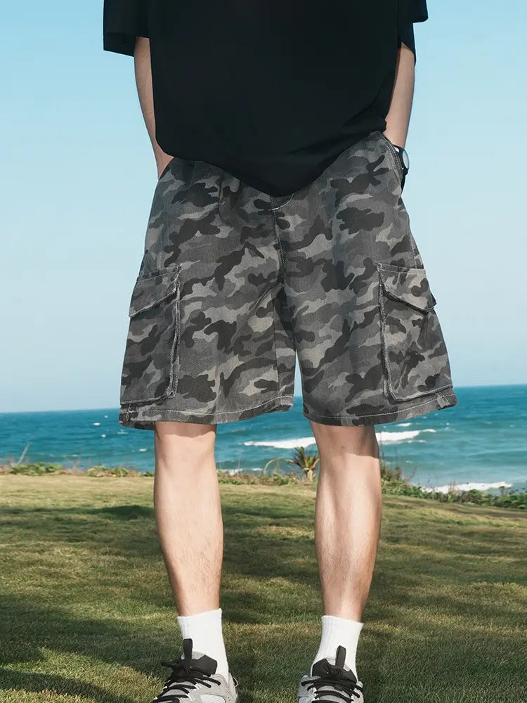 A person in Men's Dark Grey Camo Cargo Shorts with Elastic Waist and Flap Pockets, a black shirt, white socks, and sneakers stands on grass by the sea under a blue sky.