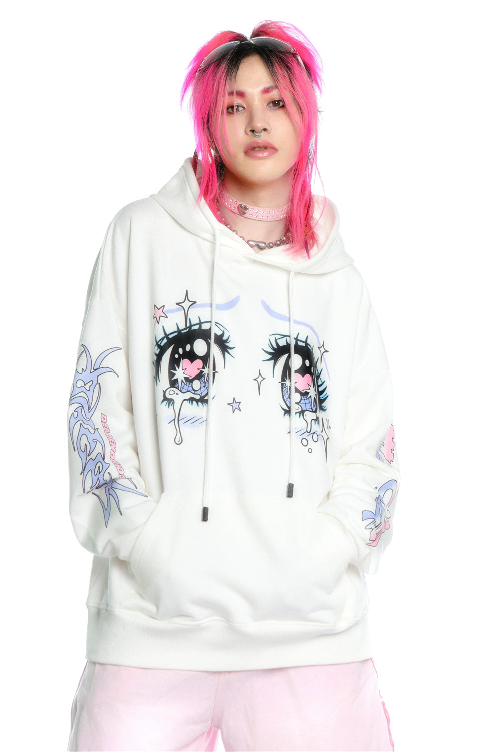 A person with bright pink hair and black roots wears the Kawaii Aesthetic Crying Eyes Hoodie by Seakoff, featuring an anime style with large cartoonish eyes and patterned sleeves. They pair it with light-colored pants and a pink choker against a plain white background.