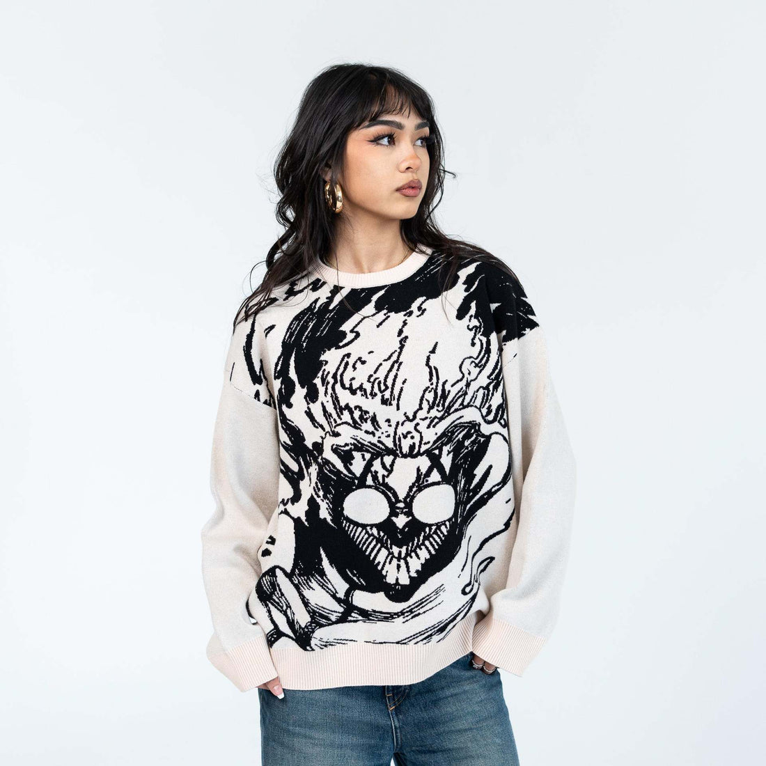 In front of a plain backdrop, someone wears the Dandadan Premium Turbo-Granny Possessed Knit Sweater, featuring a stylish black and white moth graphic. With long dark hair and hands in blue jeans pockets, they look sideways, projecting casual confidence in streetwear fashion.