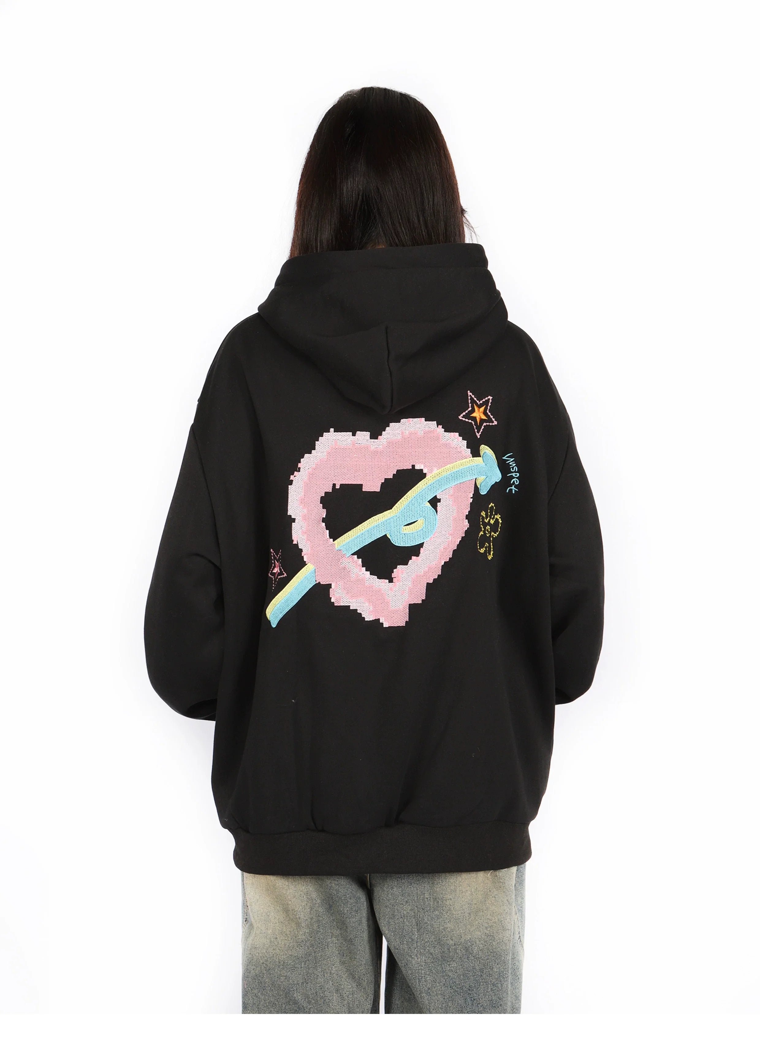 A person with long hair is wearing a Seakoff Kawaii Pastel Cartoon Hoodie featuring playful pink heart, blue ribbon, star, and flower designs on the back. Embracing kawaii fashion, they're facing away and also sporting faded jeans.