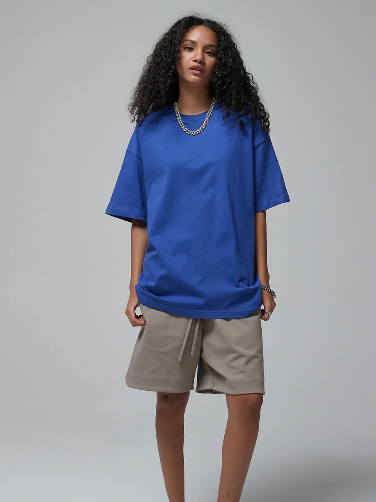 A person with long curly hair stands against a plain background wearing a Custom Anime Character Royal Blue T-Shirt—Personalized 230GSM 100% Cotton Tee, beige shorts, and silver chains.