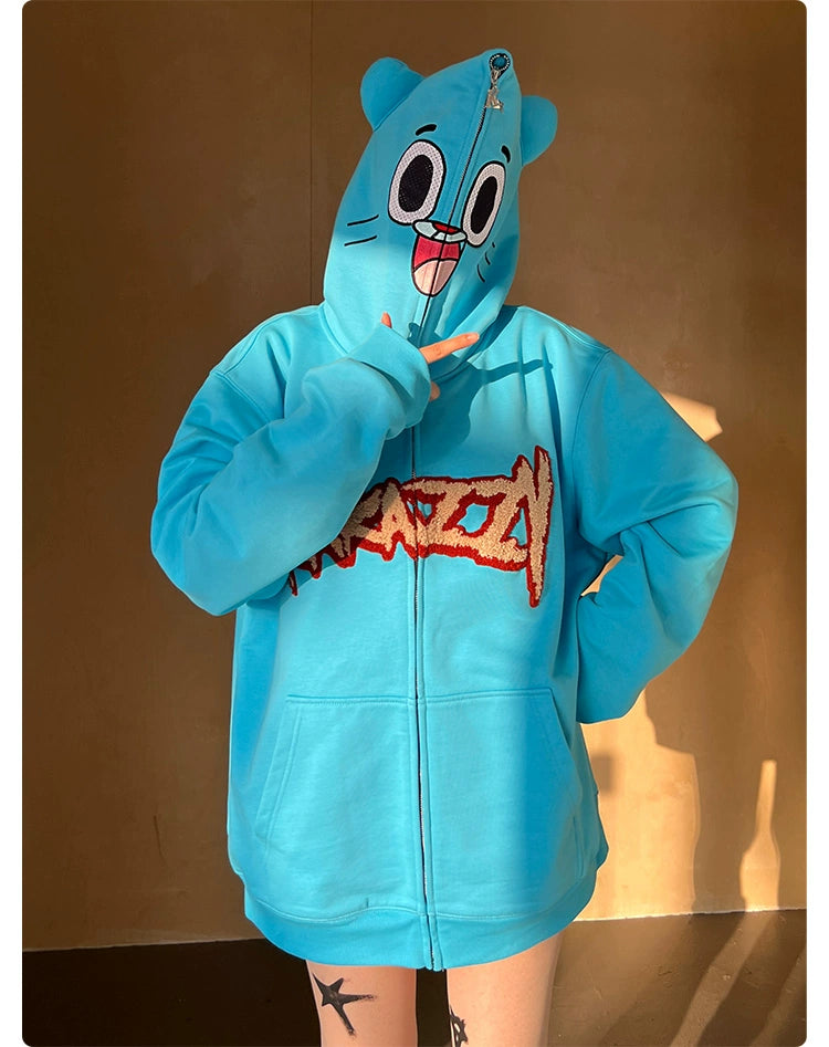 A person poses in an oversized Seakoff Adorable Blue Cartoon Hoodie, showcasing a wide-eyed face on the hood and a sturdy 400GSM cotton fabric. It features a red and white chest design, with the person making a peace sign.