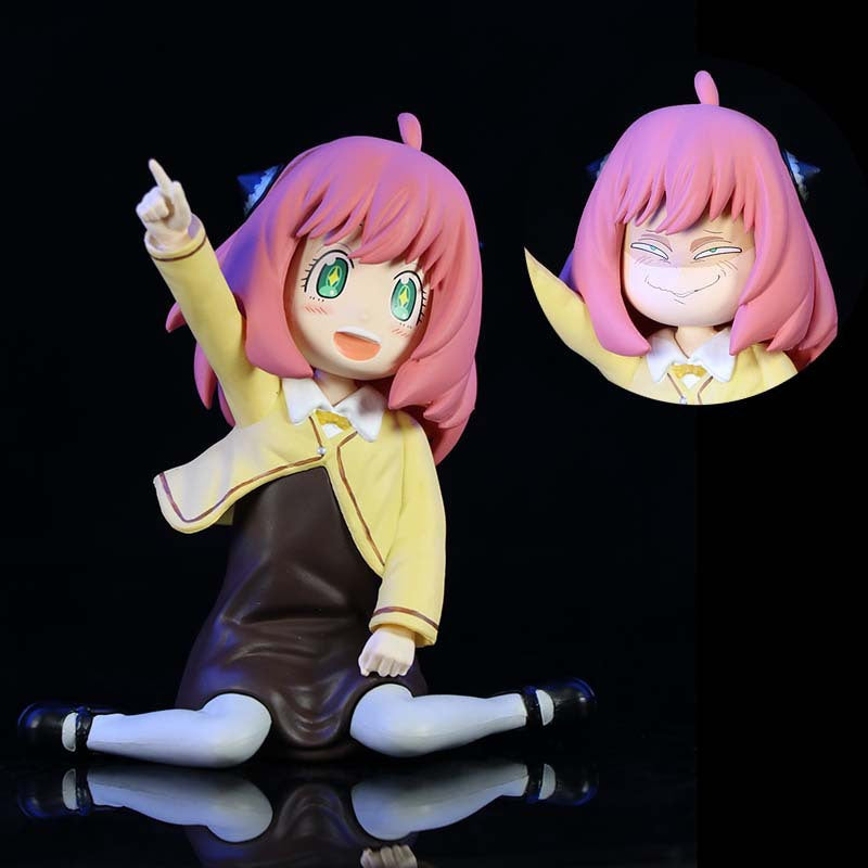 This Spy × Family collectible features Anya Forger with pink hair in a yellow school uniform over a brown dress, sitting with legs spread and pointing upward. She has large green eyes and a squinting expression.